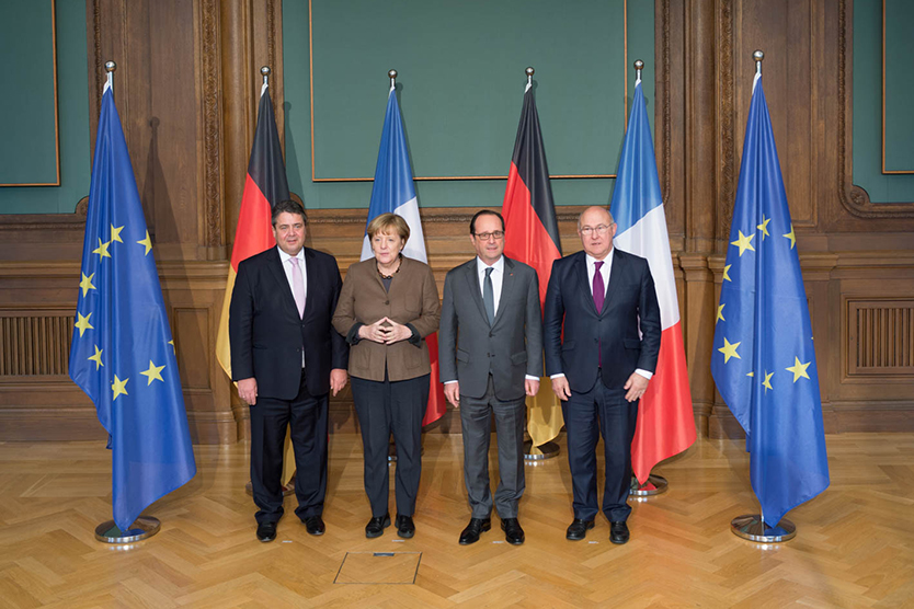 Gruppenbild mit Sigmar Gabriel, Angela Merkel, François Hollande und Michel Sapin; im Hintergund Flaggen der EU, Deutschlands und Frankreichs